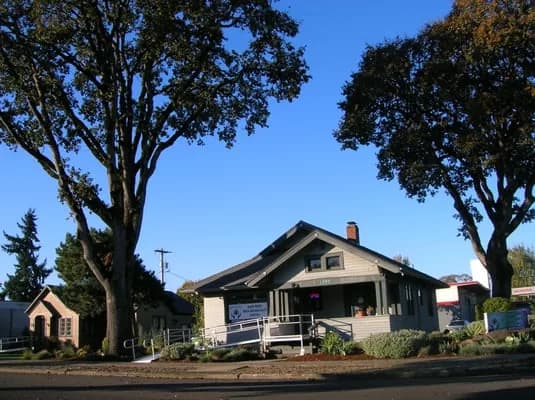 Medigreen dispensary storefront, a converted craftsman bungalow with a wraparound ramp, hipped roof, large oak trees in the front yard, and a small Medigreen signboard near the entrance.