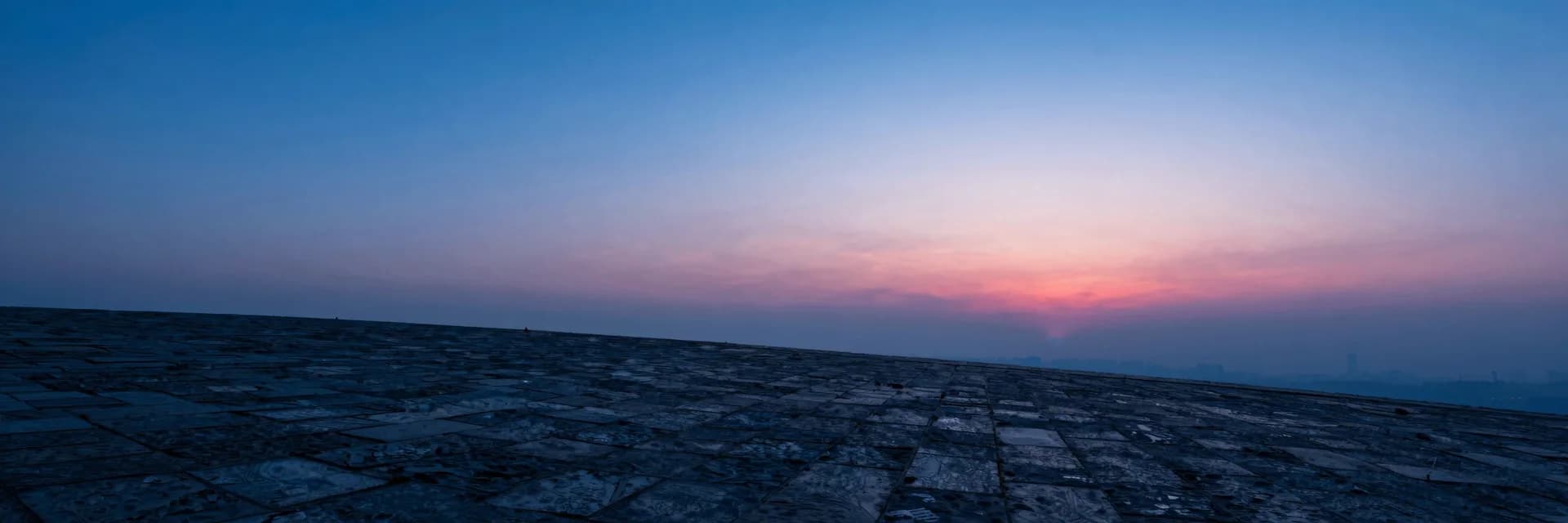 Wide atmospheric haze over a dim plain, single distant translucent monolith barely visible through electric-blue mist, hot-pink ember on the far horizon.