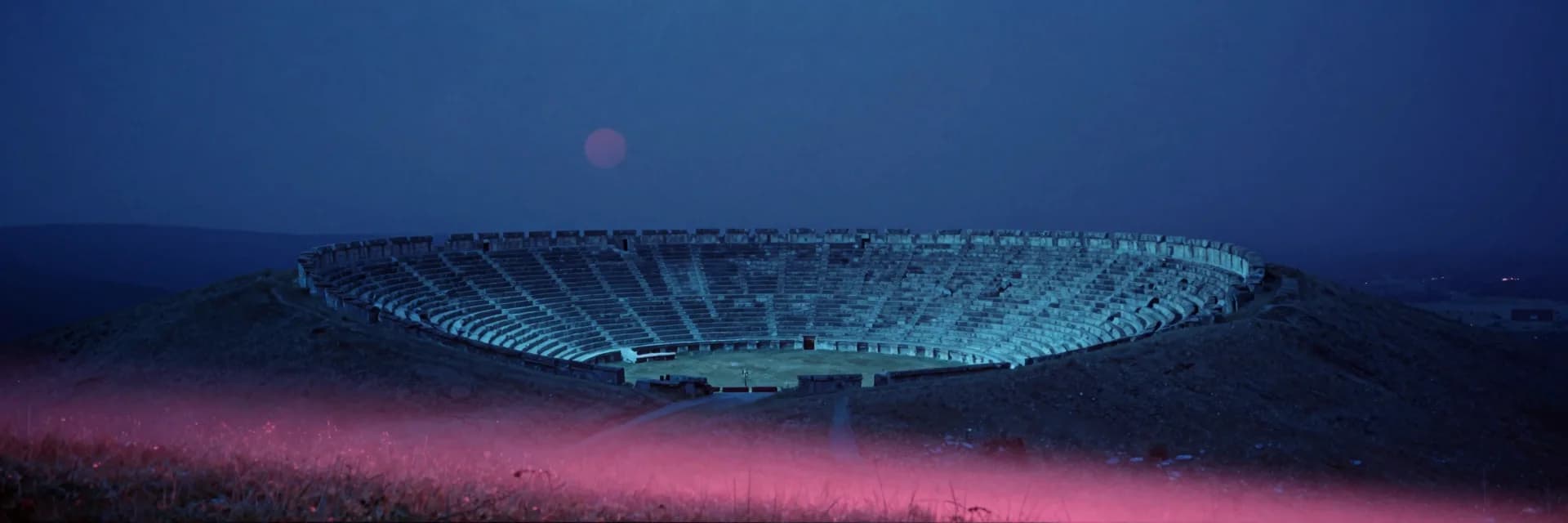 A windswept upland at dusk stretching wide, a single colossal ancient circular stone amphitheater reduced to a faint far form at the edge of frame.