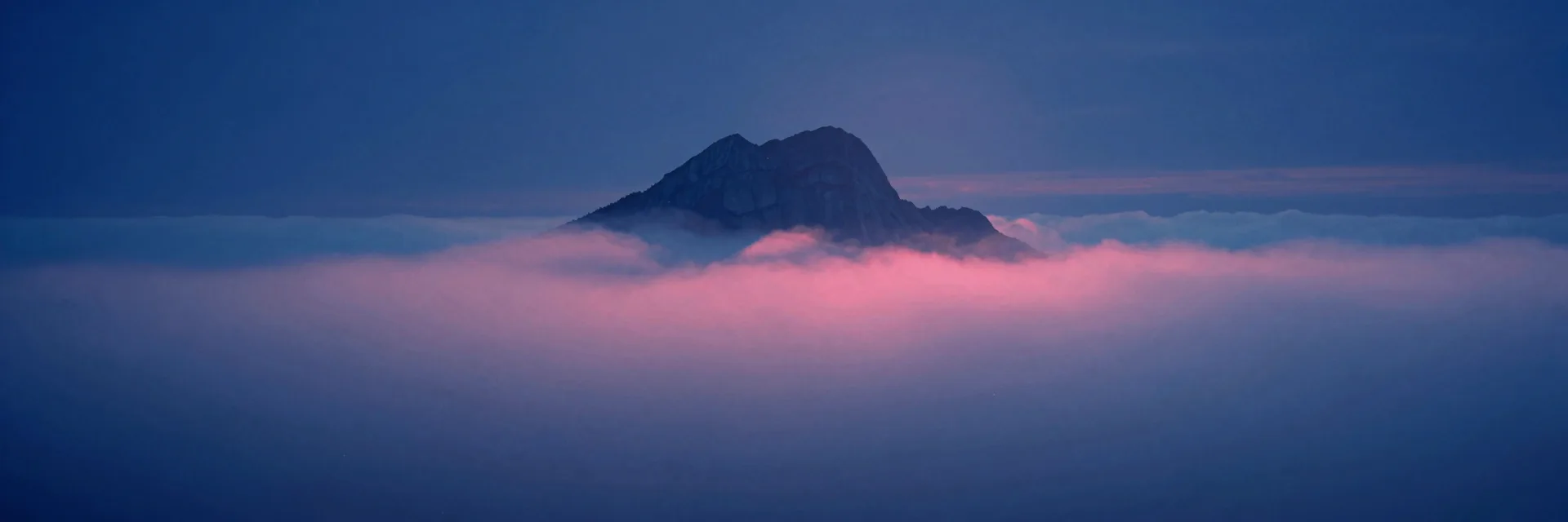 A sea of low valley fog at first dawn light stretching wide with a single broad-shouldered dolomite mountain peak reduced to a faint far form at the edge of frame.