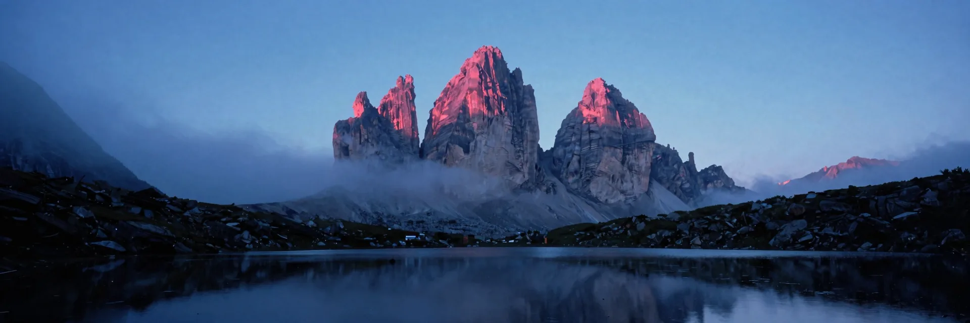 A single broad-shouldered dolomite mountain peak mirrored on a still alpine tarn at the valley floor in a sea of low valley fog at first dawn light.