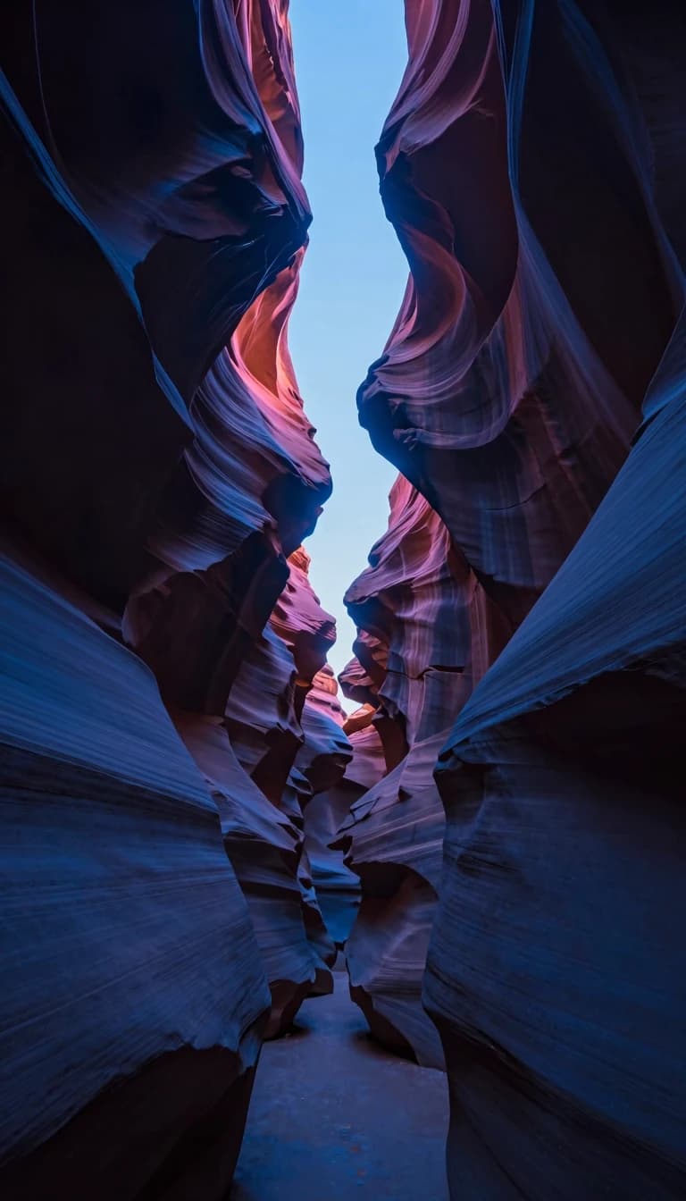 Looking up from a slot canyon floor toward a narrow strip of sky high above, canyon walls closing in from both sides, striated sandstone in deep blue shadow framing a thin band of electric-blue sky.