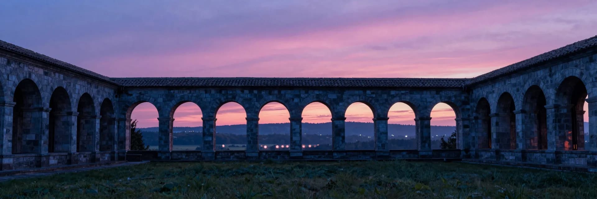Ultra-wide of the entire stone cloister structure at sunset seen from a distant vantage outside the building.