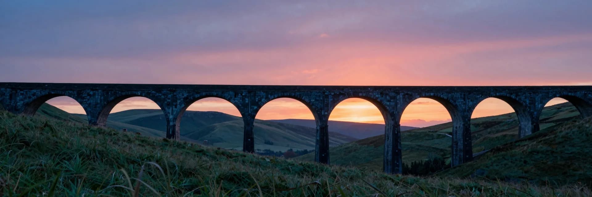 Ultra-wide of the entire stone arch bridge at sunset seen from a distant vantage, five arches visible spanning the frame, rolling green hills.