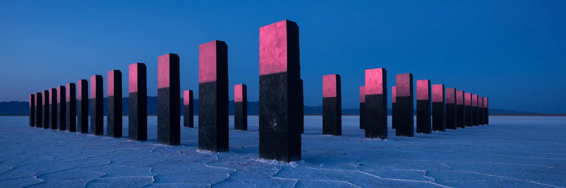 Parade-line formation of tall black monolithic slabs of equal height standing across a vast windswept salt-flat plain at twilight, the line receding into deep distance, pink alpenglow on the upper west faces of all monoliths.