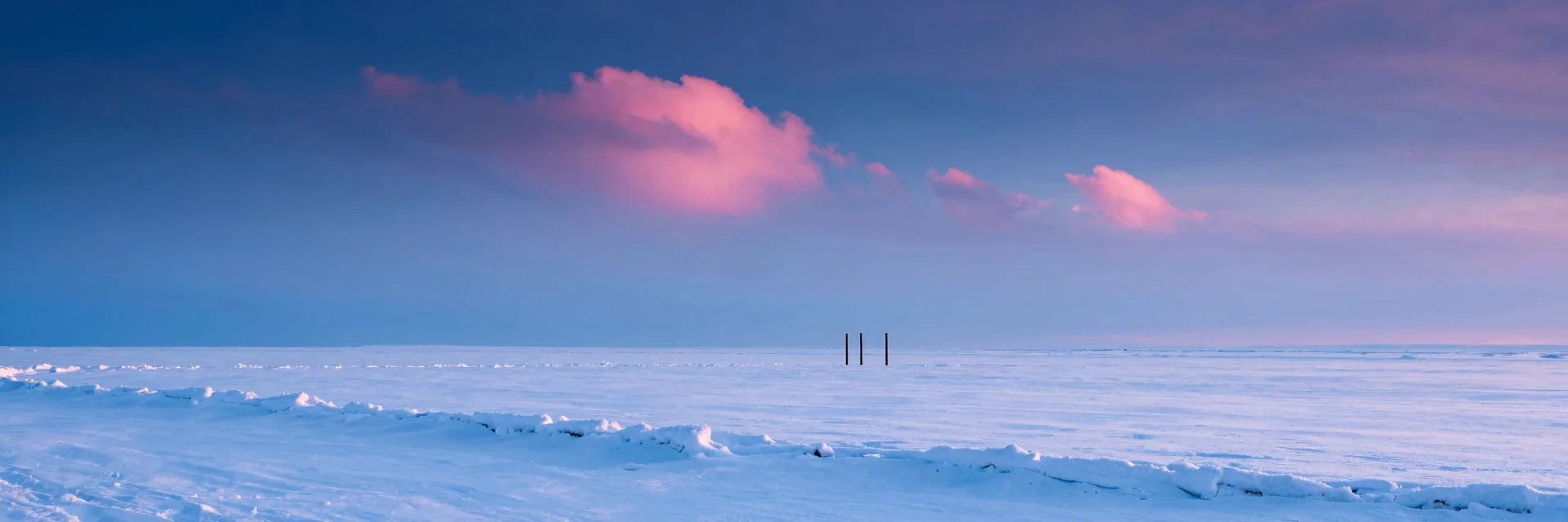 Ultra-wide of the three vertical black posts seen from a distant vantage in the vast snowy plain, dwarfed by the empty white snow and pink-clouded sky.