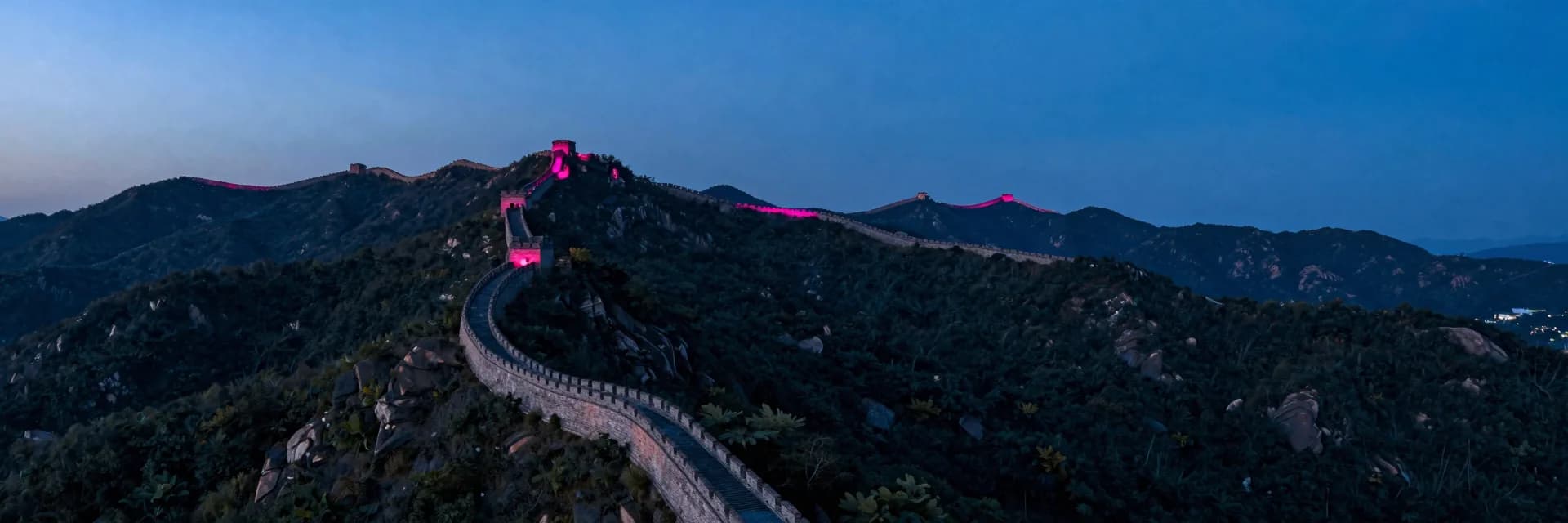 Ultra-wide aerial view of a long stone fortification wall snaking diagonally across forested mountain ridges at dusk, the wall a dark ribbon between dark forest, hot-pink sunset glow on the highest ridge crests.