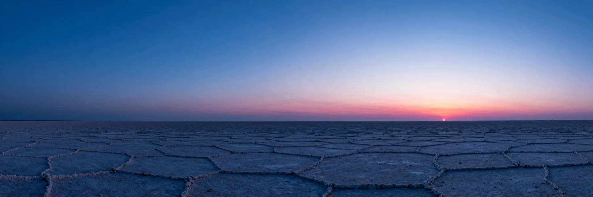 Wide atmospheric haze over a dim plain, single distant translucent monolith barely visible through electric-blue mist, hot-pink ember on the far horizon.