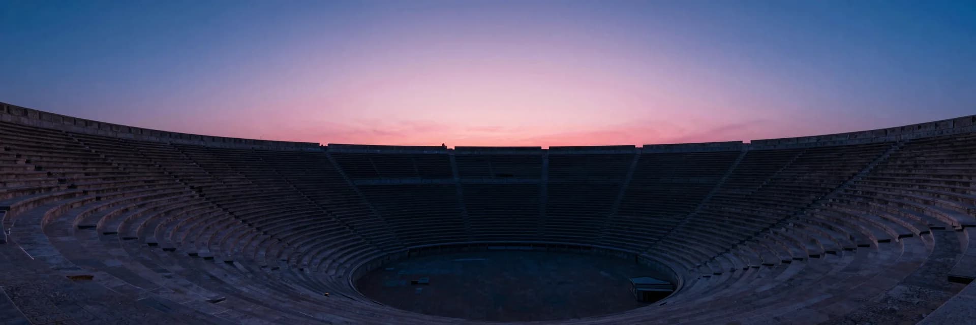An ultra-wide distant vantage from outside the amphitheater bowl showing the full curved silhouette set against a pink dusk sky.