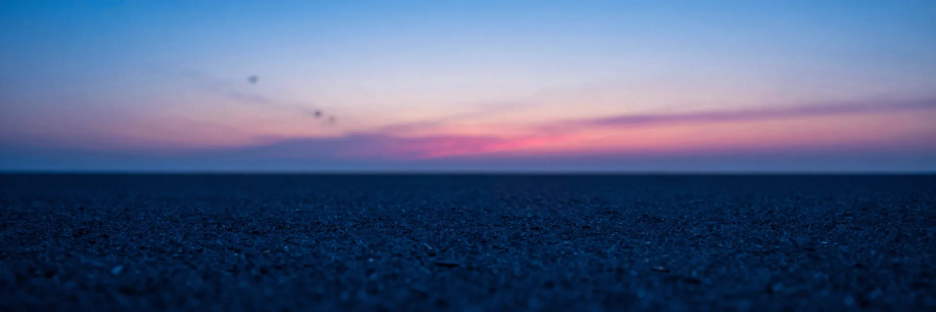 Wide atmospheric haze over a dim plain, single distant translucent monolith barely visible through electric-blue mist, hot-pink ember on the far horizon.