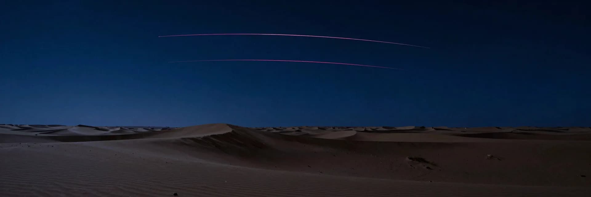 Vast desert night with three meteor streaks dwarfed by an empty dune landscape in ultra-wide format.