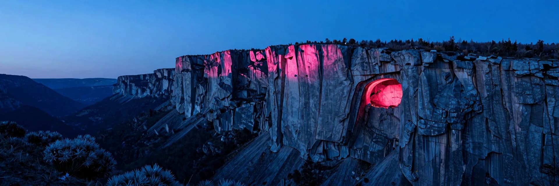 Ultra-wide view of a vast textured cliff face seen from across a deep mountain valley at dusk, single pinkish-red glowing cave opening visible small in the right third, dwarfed by the cliff scale.