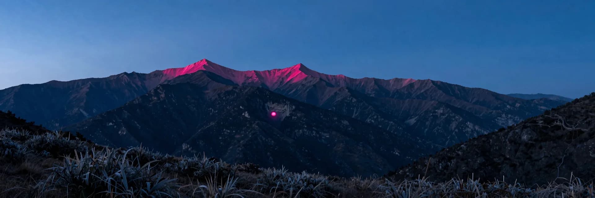 Ultra-wide layered mountain silhouettes at dusk with the single pinkish-red glowing cave visible as a tiny pink point on a cliff face in the middle distance, dwarfed by the mountain scale.