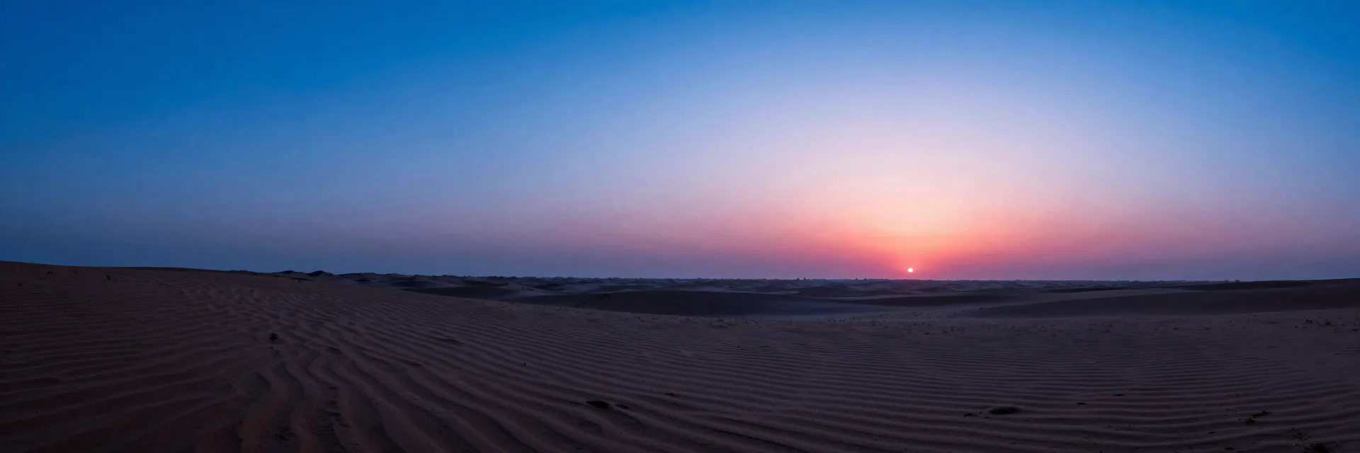 Wide atmospheric landscape at dusk, deep electric blue sky over a quiet plain with a low pink horizon glow softening the foreground.