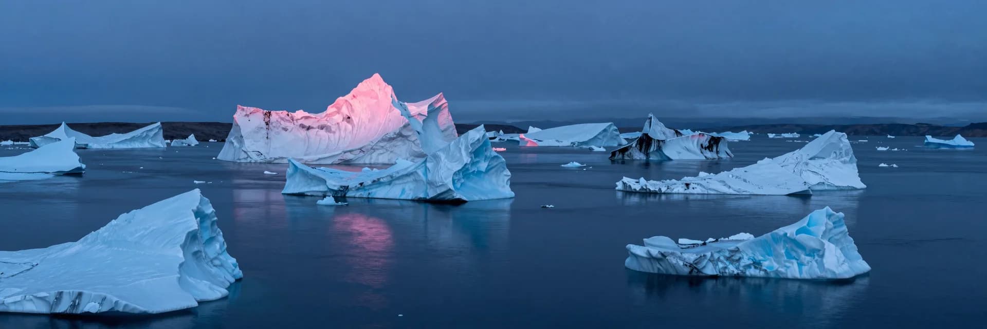 Ultra-wide aerial view at dusk over partially-frozen water with multiple ice formations breaking the surface, hot-pink dusk glow on the highest spire tops.