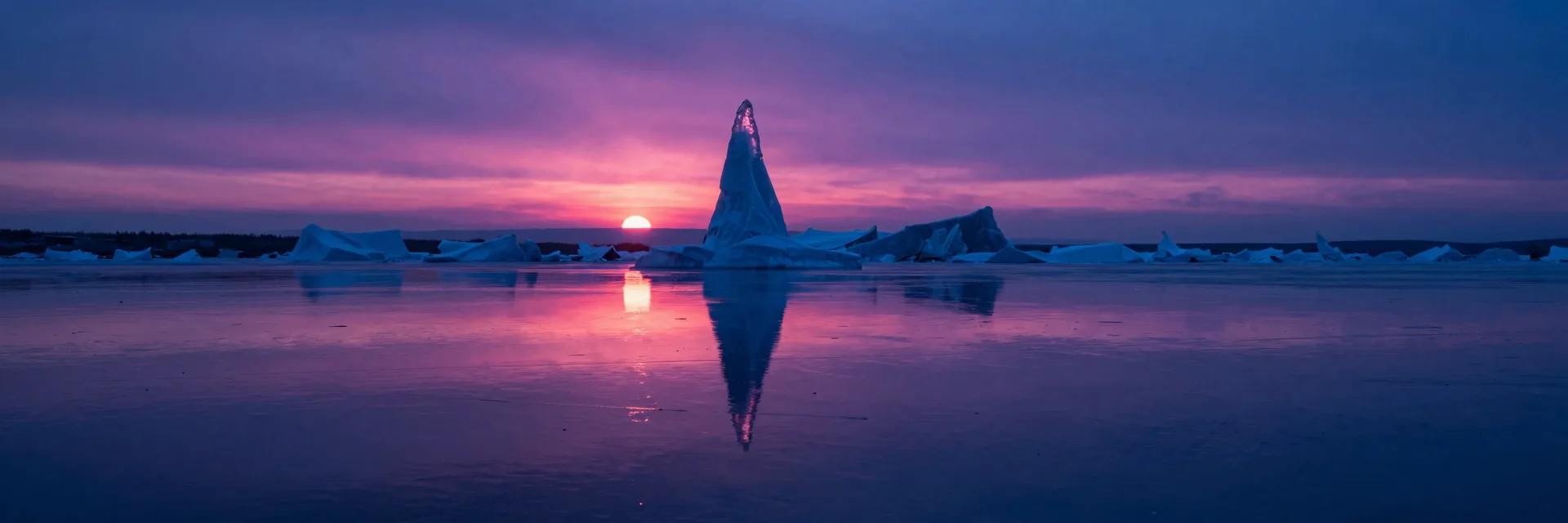Ultra-wide view of a single ice spire reflected on a perfectly still partially-frozen water surface at sunset, mirror composition with reflection extending downward, hot-pink sunset glow on the spire top and reflected on the water.