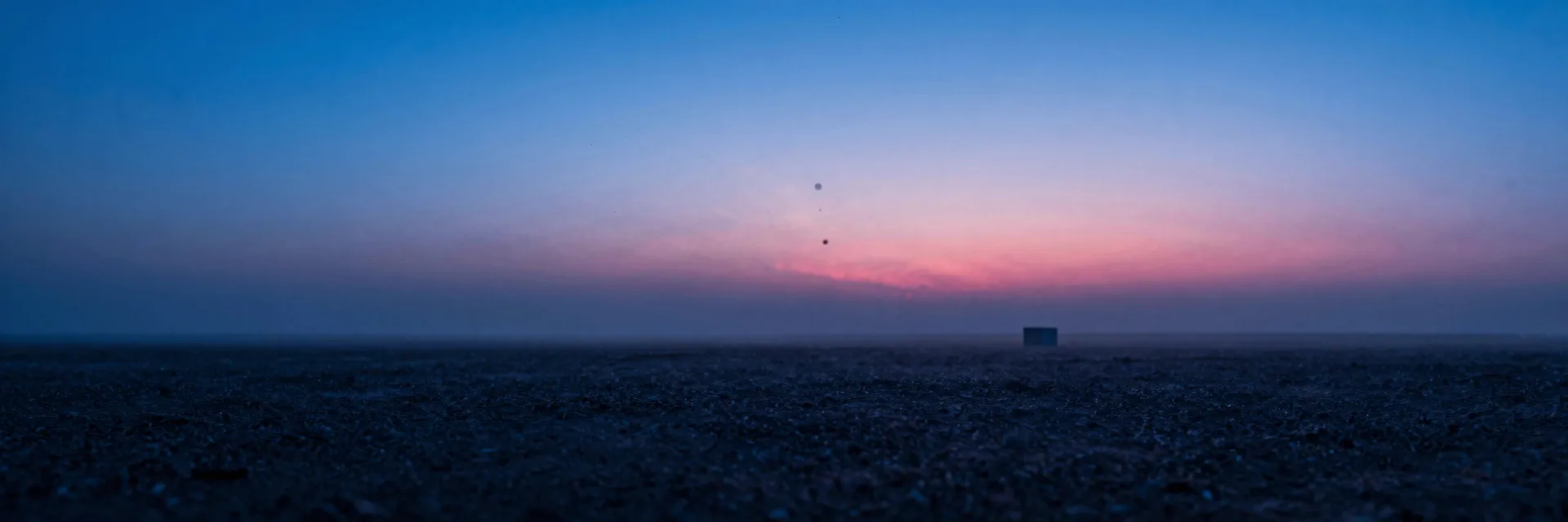 Wide atmospheric landscape at dusk, deep electric blue sky over a quiet plain with a low pink horizon glow softening the foreground.