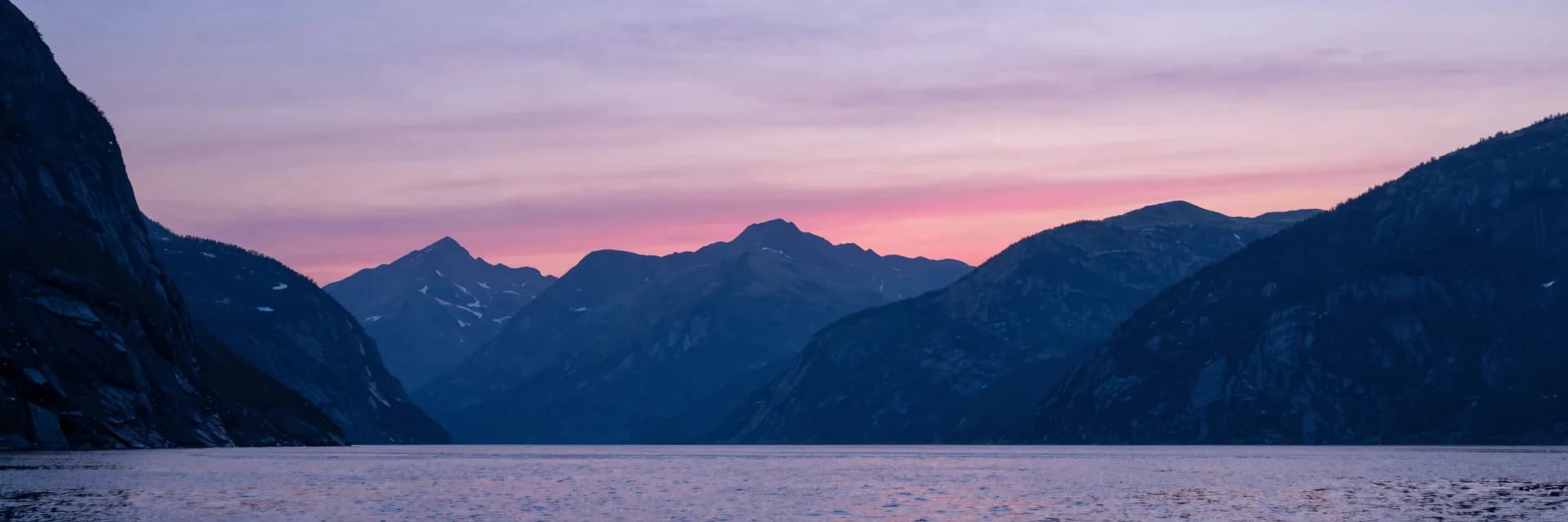 Ultra-wide of the distant slightly-blurred mountain ranges visible past the fjord opening, layered mountains receding under the pink sky.