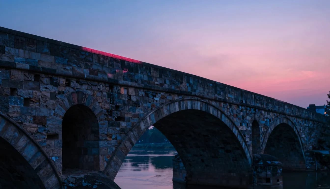 Side view of an ancient stone arch bridge from along the riverbank with the sun setting beyond it.