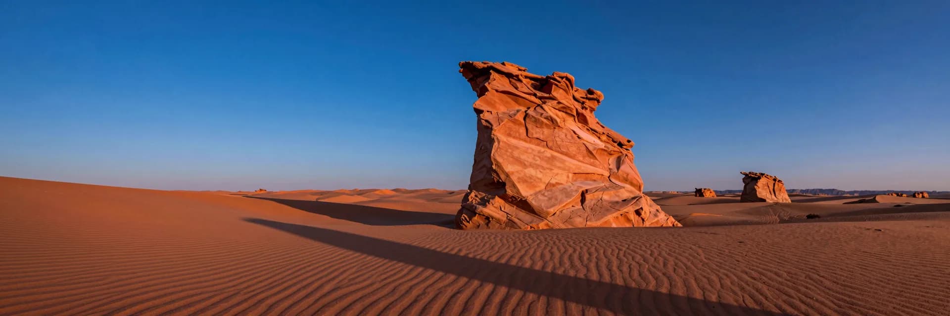 Ultra-wide of the twisted hoodoo at low golden-hour sun casting a long dramatic shadow across the rippled orange desert floor toward the foreground.