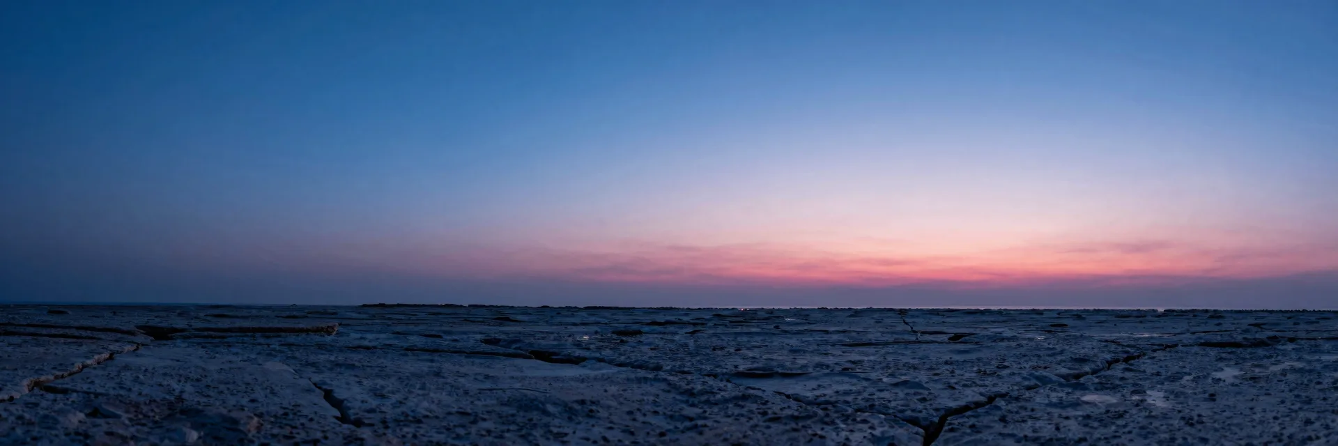Wide atmospheric haze over a dim plain, single distant translucent monolith barely visible through electric-blue mist, hot-pink ember on the far horizon.
