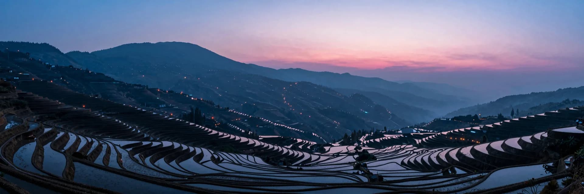 Ultra-wide of the vast terraced hillside at dusk seen from a distant elevated vantage, wavy patterns of paddies cascading, layers of misty hills.