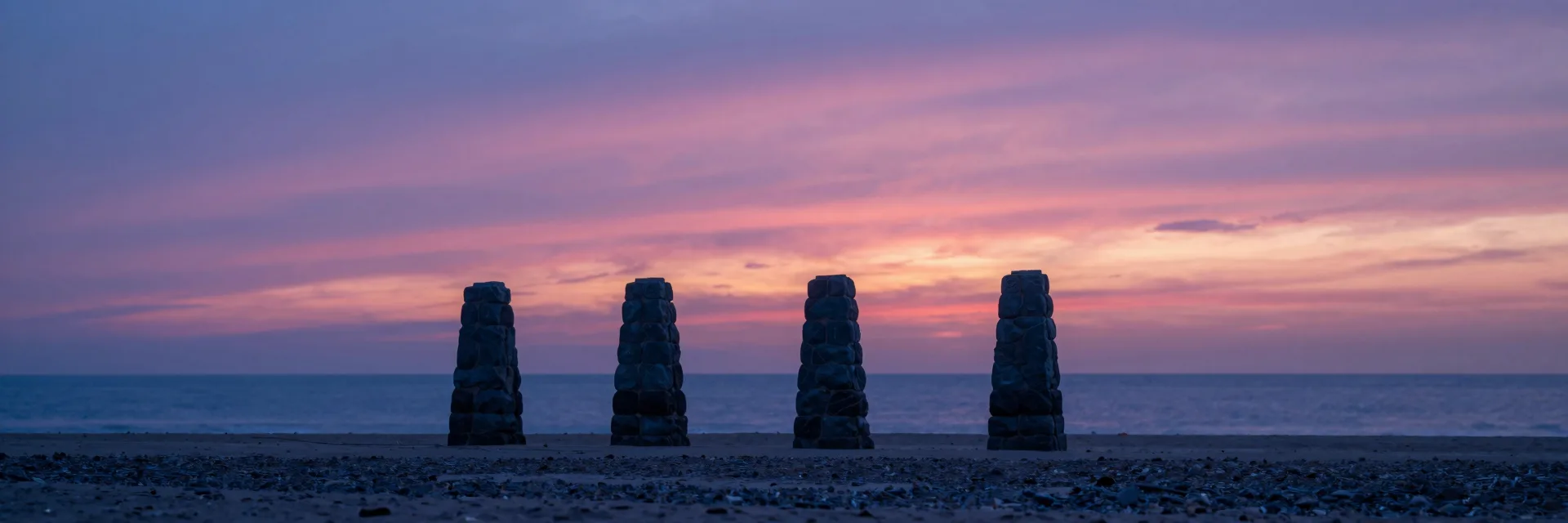 Ultra-wide of the four stone pillars at sunset seen from a distant vantage along the coast, all four pillars visible as a row in the ocean.