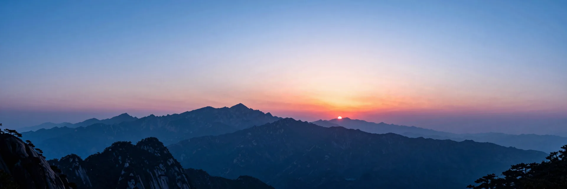 Ultra-wide of multiple layered mountain ranges silhouetted at sunrise, each layer progressively lighter as it recedes into atmospheric haze, the sun rising behind the highest peaks.