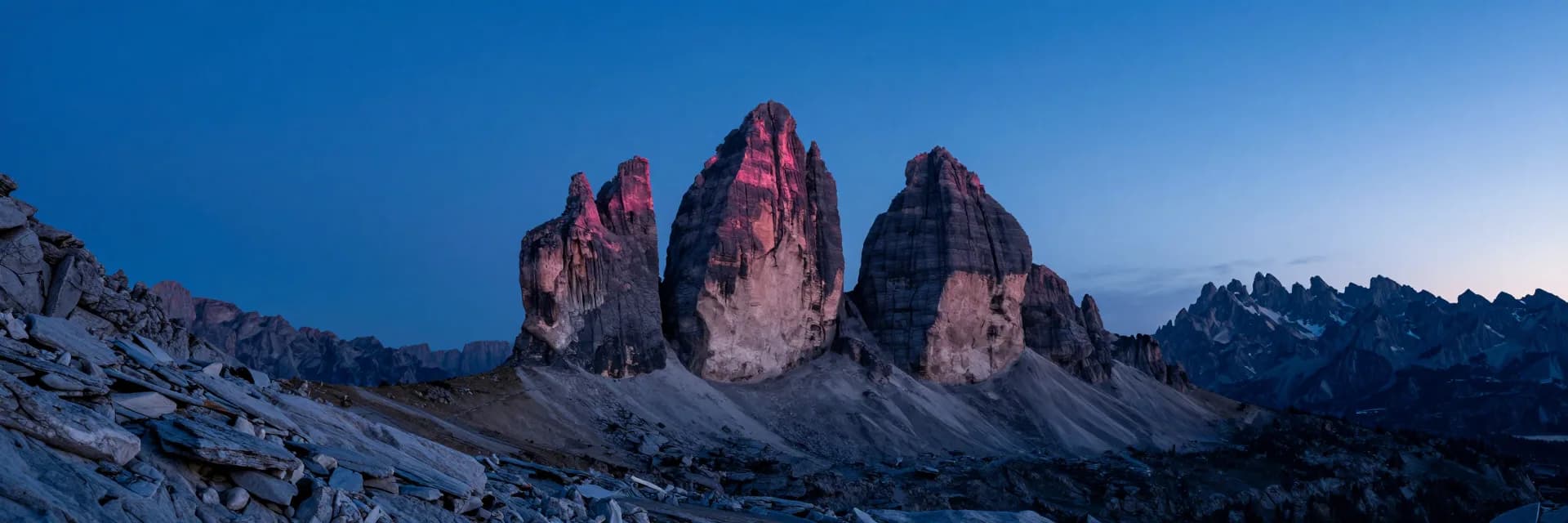 Cinematic ultra-wide panorama of dolomite mountain ridges receding into deep distance, three sharp spires centered, pink alpenglow on the highest crests.