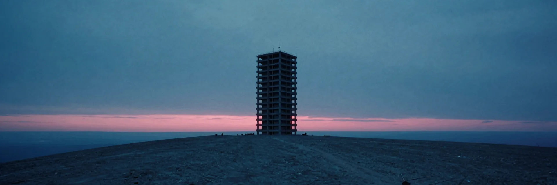 An ultra-wide panorama of a single brutalist tower standing alone on the crest of a low rise, a pink dusk band glowing along the horizon behind it.