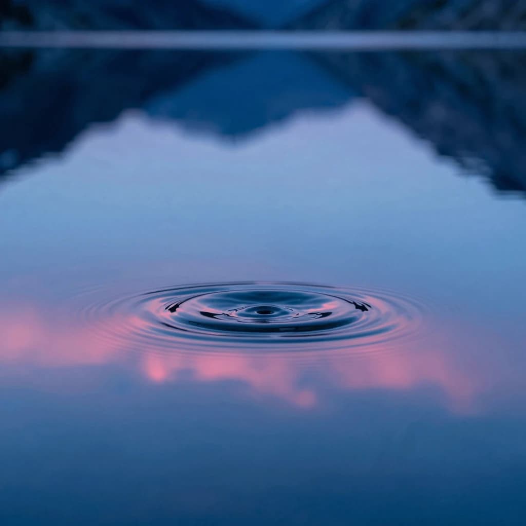 Macro of an alpine tarn water surface with a single concentric ripple expanding outward, glassy mirror-still water surrounding the ripple, electric-blue dawn sky reflecting around it.