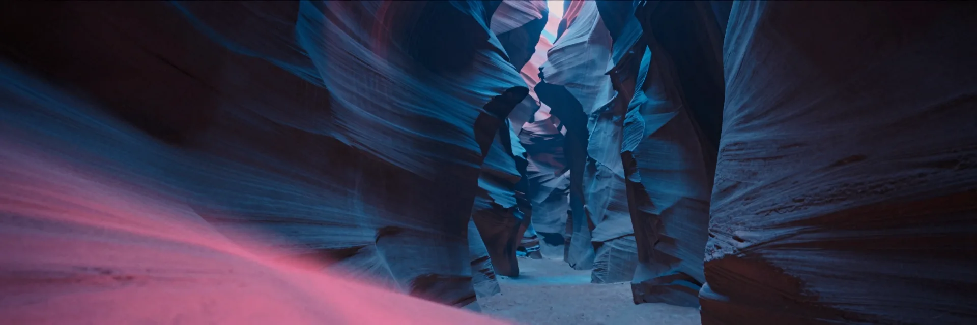 A deep slot canyon stretching wide, a single immense vertical canyon wall split down the center by a hairline fissure of light reduced to a faint far form at the edge of frame.