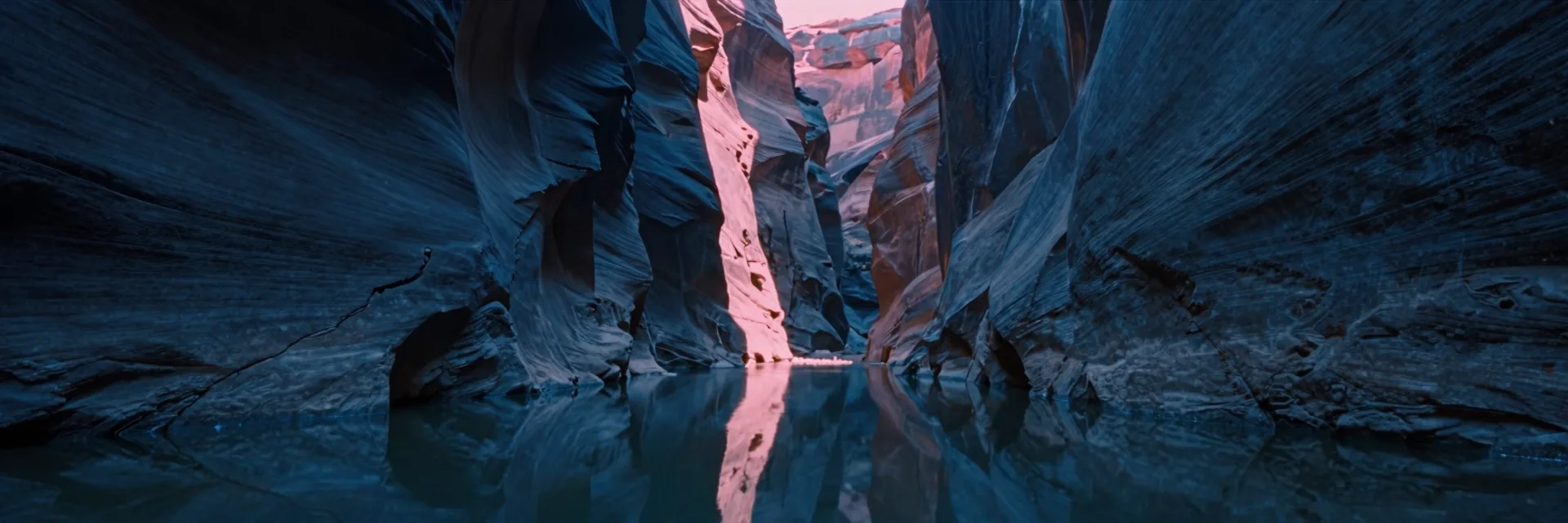 A single immense vertical canyon wall split down the center by a hairline fissure of light mirrored on a still pool at the canyon floor in a deep slot canyon.