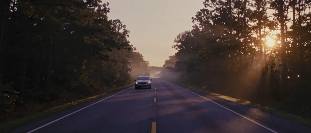 A lone car on a rural two-lane road at dawn, pines on both sides.