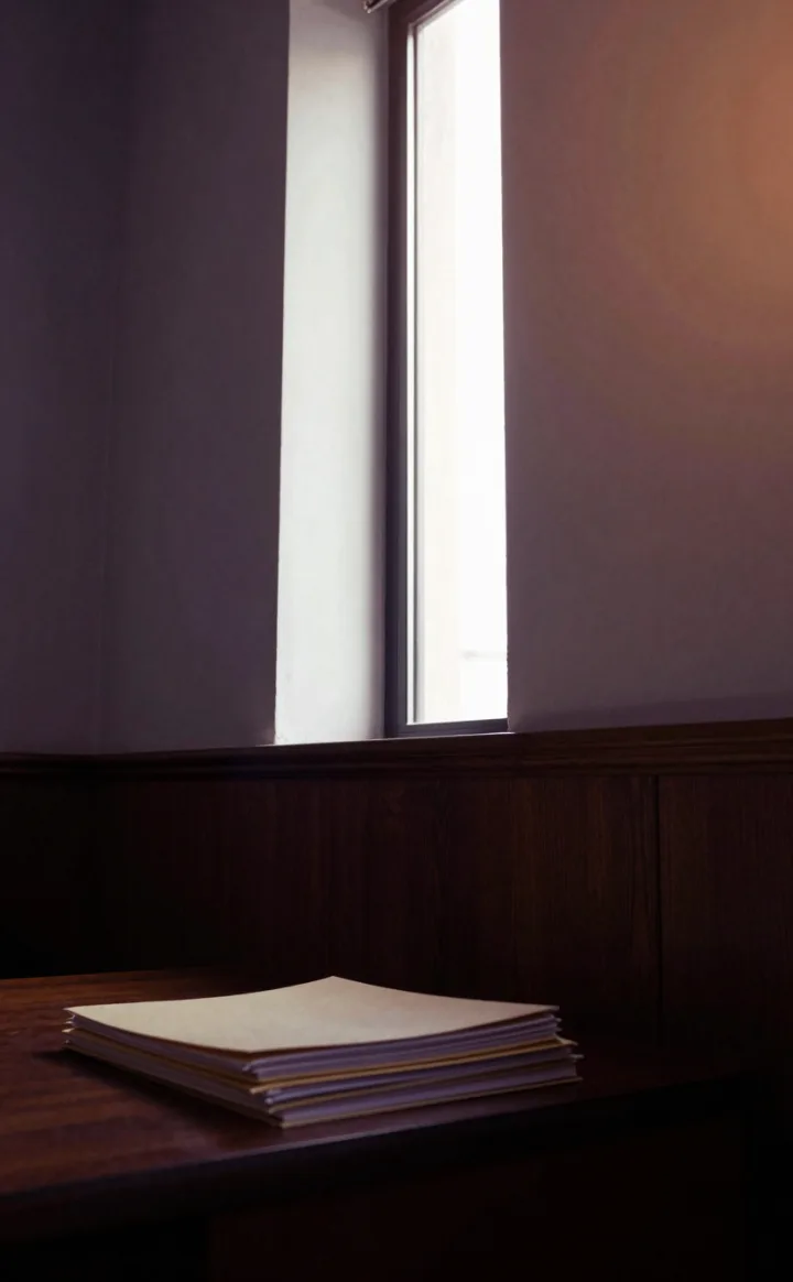 A desk corner by a window with a stack of papers.